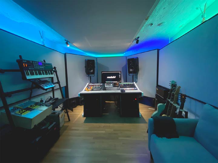 Nadoki studio, at Riverside Studios (Berlin), being lit by blue light. On the left, there is a stand with MIDI keyboards and synthesizers. In the middle, there's a black office chair facing a black studio desk equipped with a computer screen, outboard gear, and two pairs of speakers. On the right, there are two guitars on a stand and a guitar amplifier. The studio walls and ceiling are full of acoustic panels.