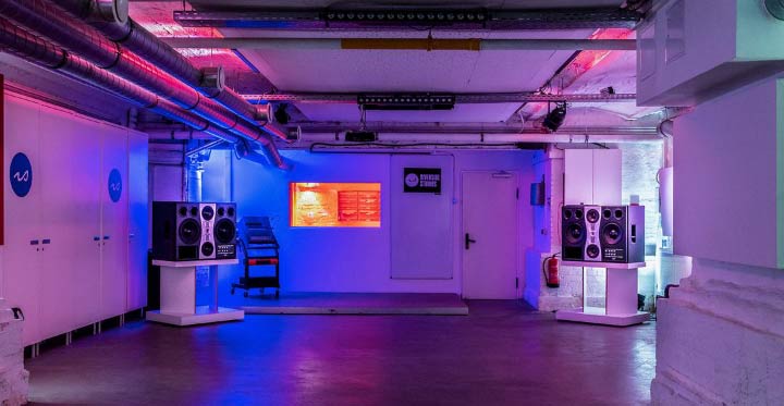 The Lab, at Riverside Studios (Berlin), being lit by blue and red lights. The ceiling is full of pipes and cables, there's a a collumn on the right and, on the back, there's a pair of big speakers and a door. Most of the room is empty.